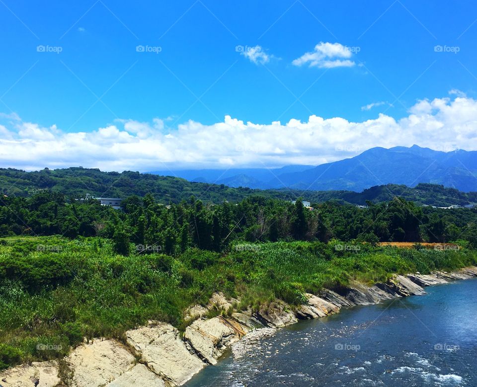 A beautiful green landscape full of trees. In the foreground is a steady stream. In the background are mountains and a deep blue sky.