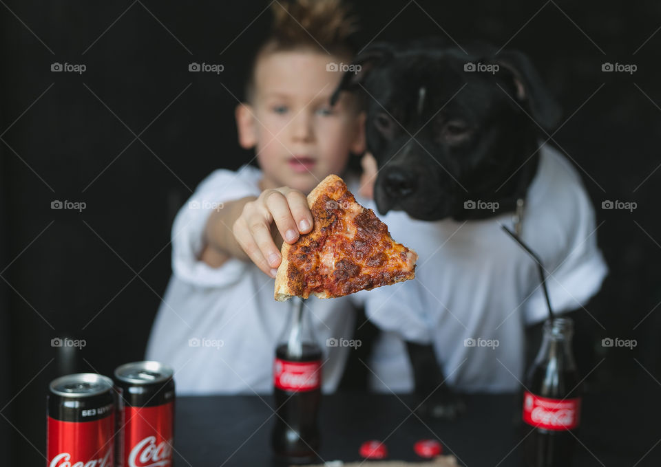 cute kid with his friend dog, dressed in white T-shirts, sitting at the table, eating pizza and drinking Coca-Cola.  Emotions, joy.  Black background