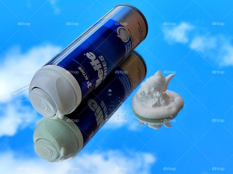 On the mirror surface lies a blue bottle with a white inscription: "Gillette series" and white fluffy foam. Objects and a blue sky with white clouds are reflected in the mirror surface