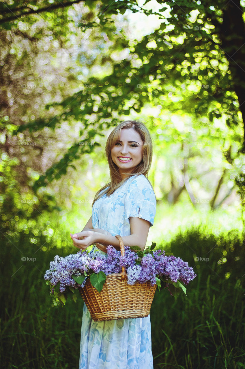 Portrait of beautiful young smiling lady in blue dress with basket of purple lilacs. Spring colors and backgrounds.