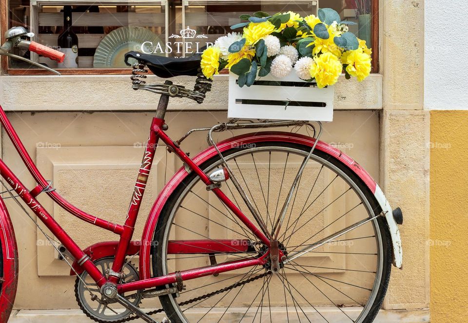 A red bicycle with a box of yellow flowers on its rear, leaned up in front of a shop.