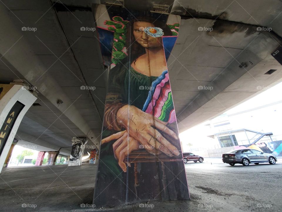 Visual street art on cement Highway pillar under highway in a parking lot. Appears to be text-mix inspired Mona Lisa.