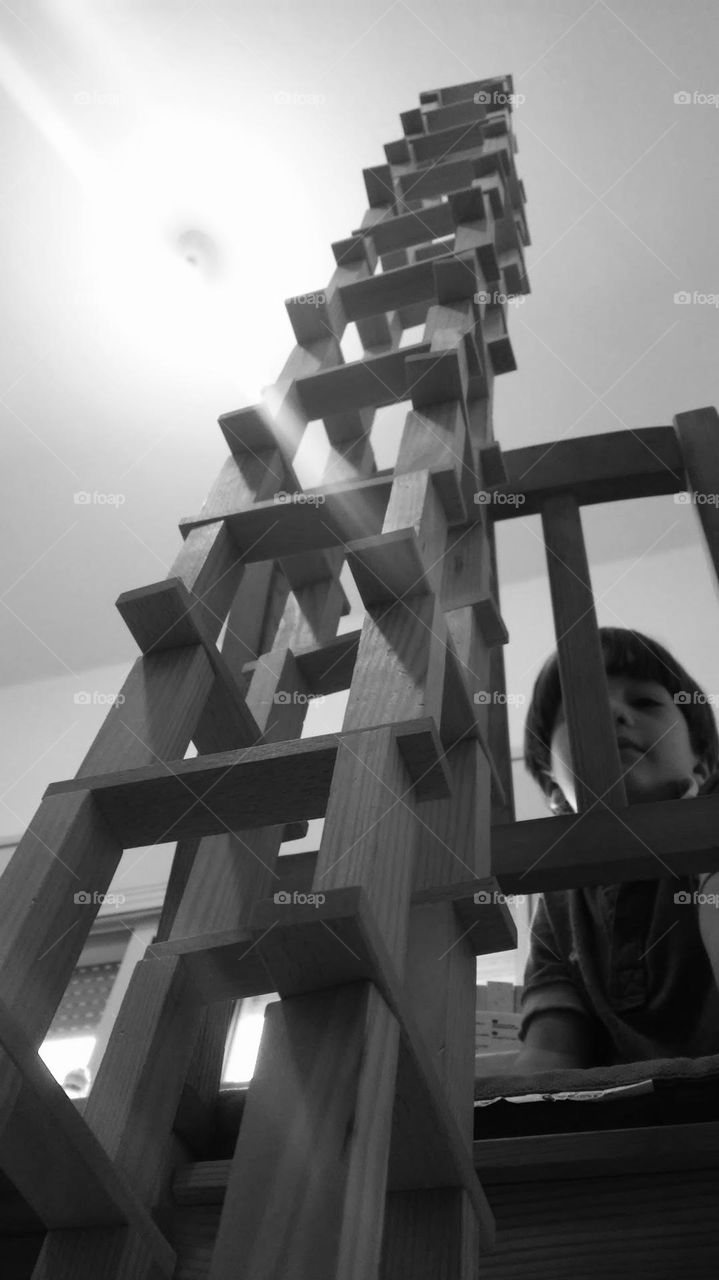 Very high tower made of wooden blocks with kid peeking while climbing chair for continuo building