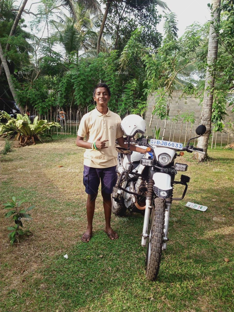 This is a image of a teenager and his motorcycle. He is in happy mood and the motorcycle is seems like a racing cycle. He is standing on grass and the surrounding is green in colour because of vegetation .