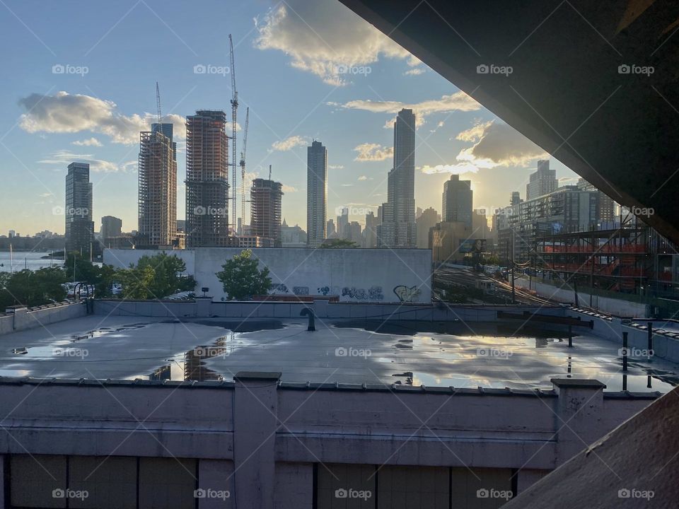 “Long Island City” and in the distance “Manhattan” can be seen from here descending the staircase of the “Pulaski Bridge”. The setting sun adds a beautiful warm golden light and reflects off wet areas on the rooftop across. 2023. Hypnotic Productions