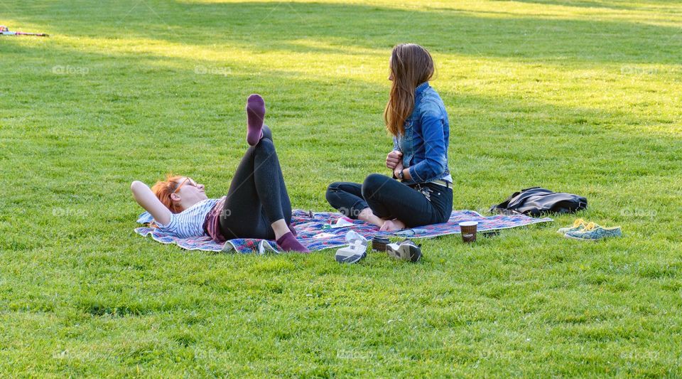 Two female friends enjoying and relaxing on blanket in park in city