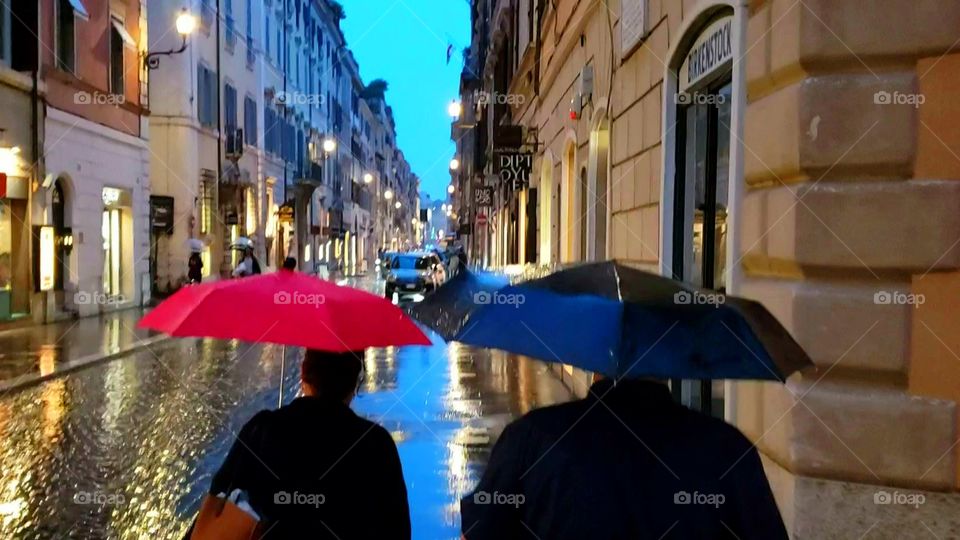 Two people walking in the rain, with red and blue umbrellas, down a street in Rome, Italy, during evening with streetlights reflecting on the side walk. Romantic.