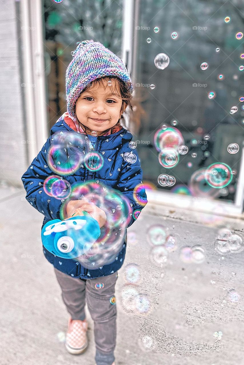 Child blowing bubbles outside in the winter, Circles outside in the form of bubbles, circle shaped bubbles, toddler loves bubbles, toddler having fun with bubbles, outside with a bubble maker, running with bubbles surrounding toddler, fun outside 