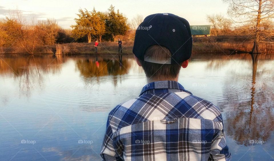 Boy fishing a Pond in Fall with two other boys across the bank from him fishing in the distance