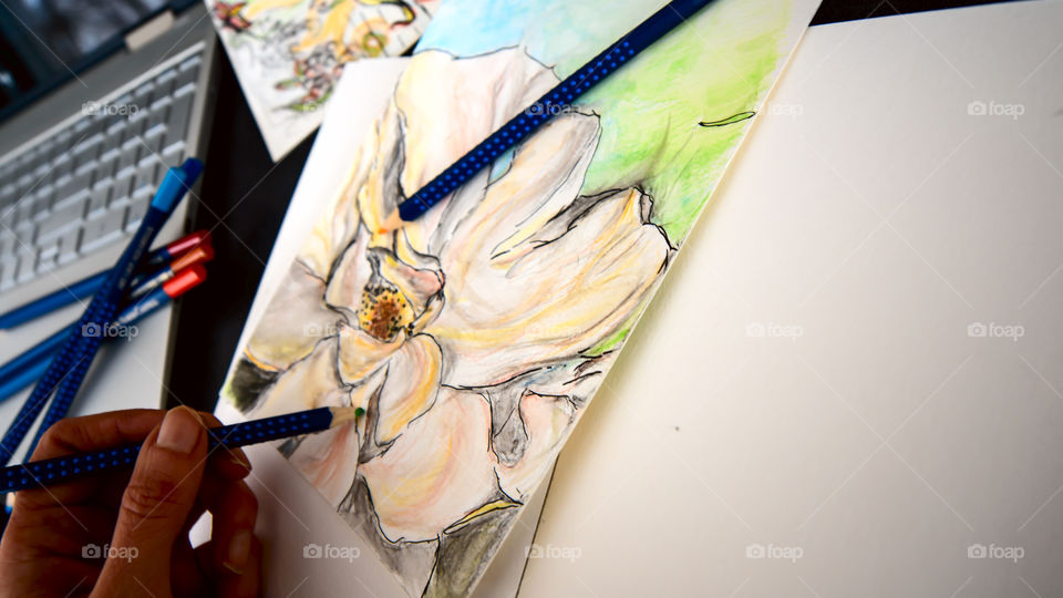 Closeup of hand holding Faber-Castell Aquarelle Colored watercolor pencils sketching closeup spring flower with sketchbook on table with laptop and coffee conceptual work life balance, creativity, hobby artist lifestyle photography