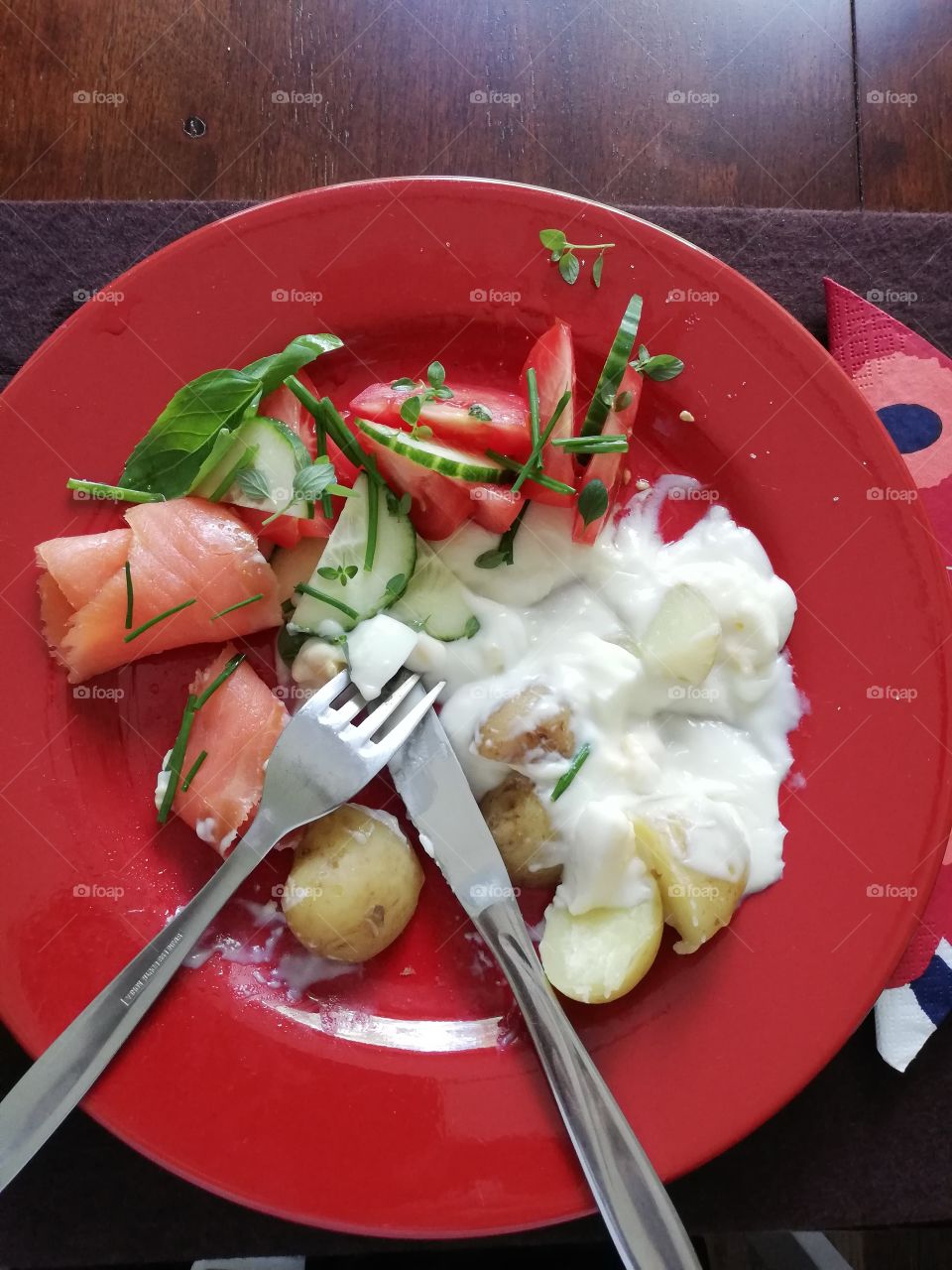 Fresh hot meal on a red round plate on a brown wooden table. Rolled cold smoked salmon, boiled eggs in white sauce and potatoes with skin. Cut cucumber and tomato, basil, thyme, chives. Fork on the knife, a napkin under the plate.