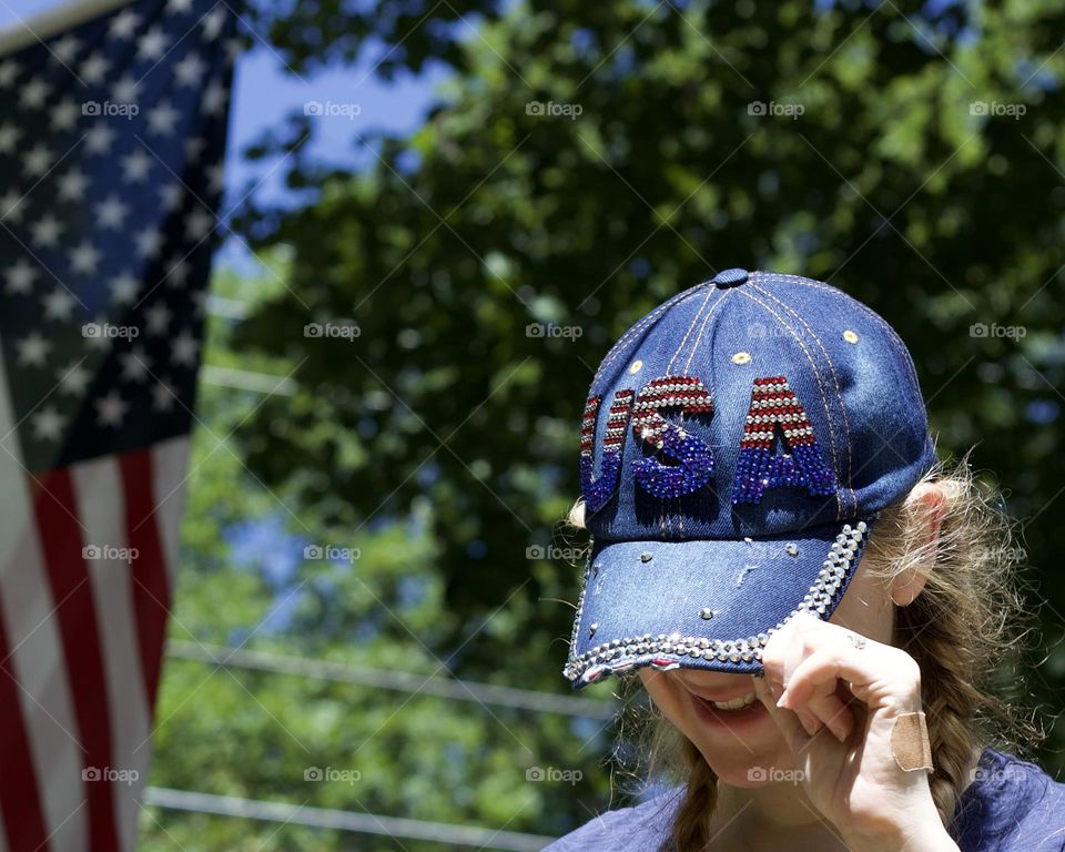 Born in the USA; Woman sporting a Bedazzles USA baseball cap with trees and American flag in the background 