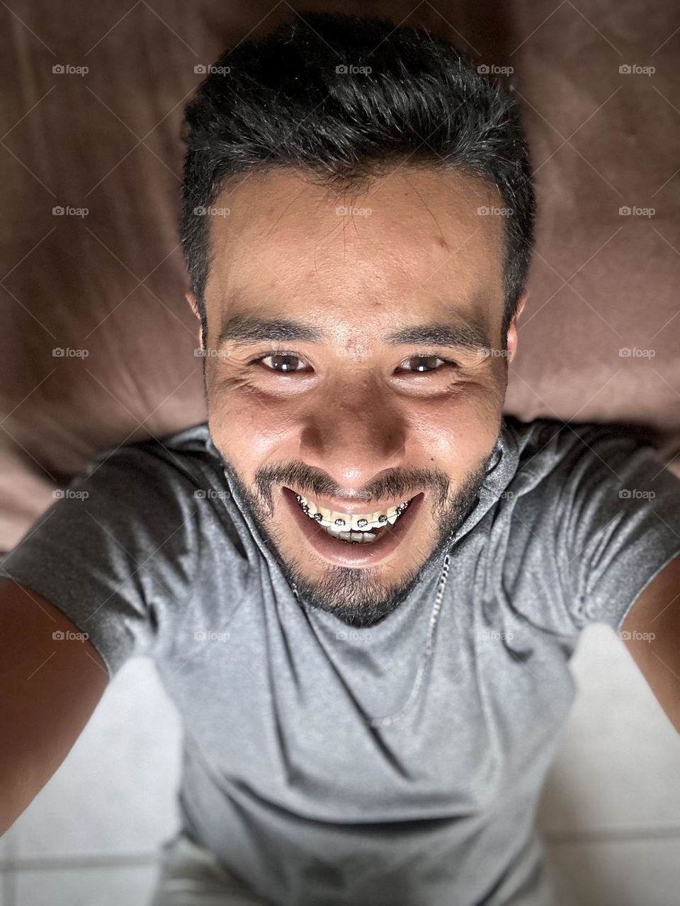 Man lying down on his bed while he looks to the camera and laughing and looking happy at his face.