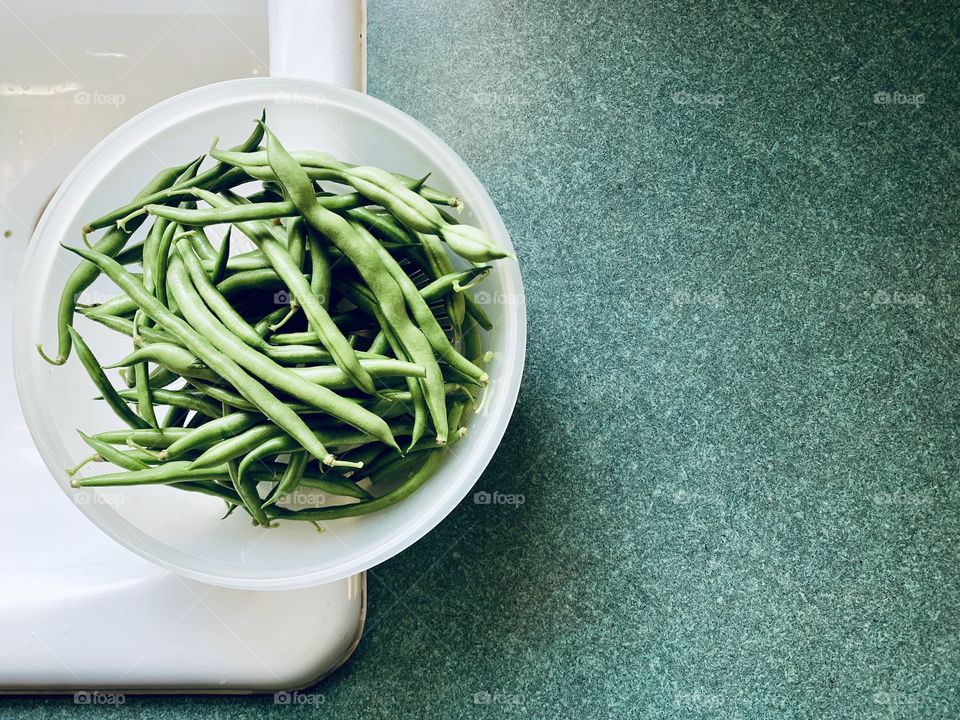 Overhead view of garden-fresh green beans in a white plastic colander on the edge of a white sink by a green kitchen countertop