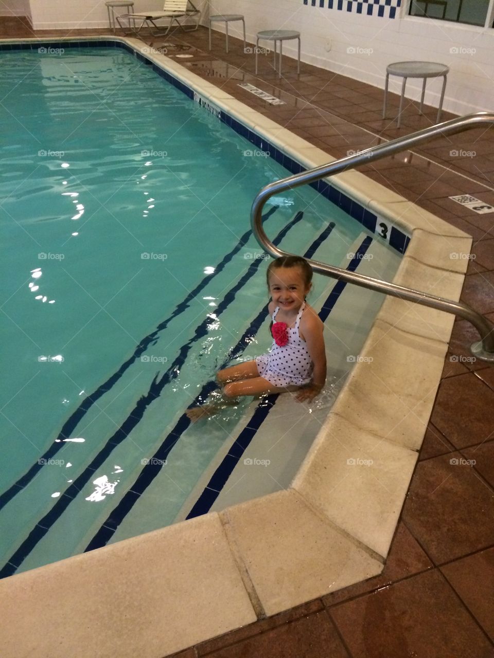 Poolside Cutie. Little girl in retro style bathing suit and braids sitting on the edge of an indoor pool, smiling. 