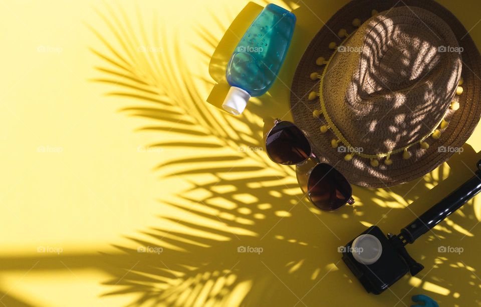 Straw hat, camera, sunblock and sunglasses lie to the right on a delta background with copy space on the left with palm tree shadow, flat lay close up. Concept summer, blogger, vacation, travel.