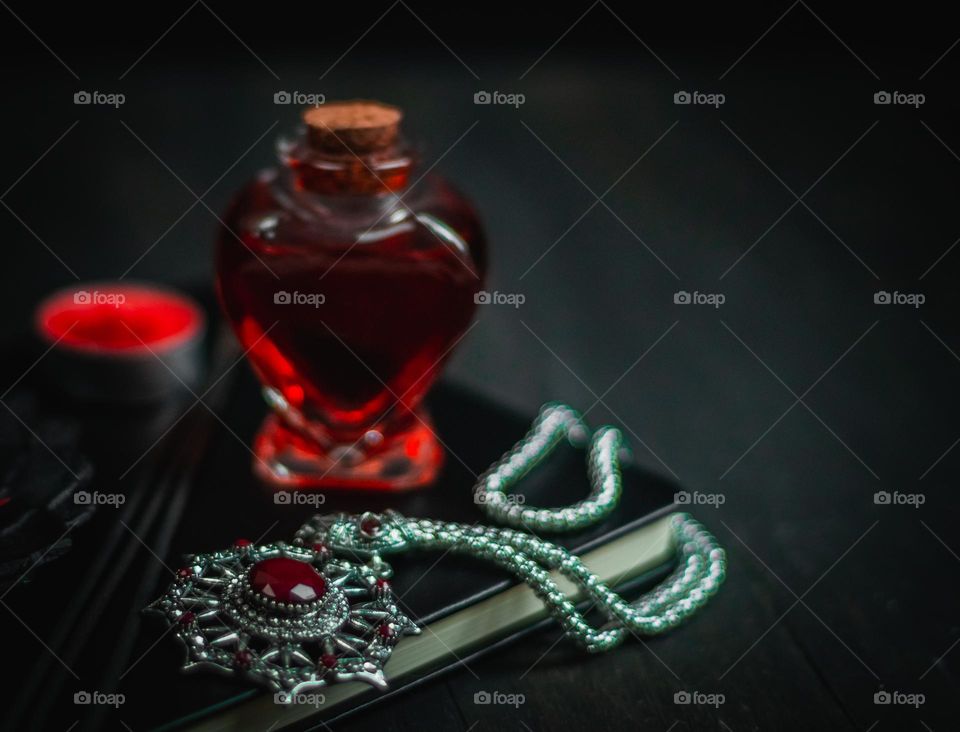 One vintage amulet with red stones on a silver chain sits on a black book with a heart-shaped red vial of poison and a burning candle on a black ion, close-up side view. The concept of ancient jewelry, vintage amulet, esoteric.