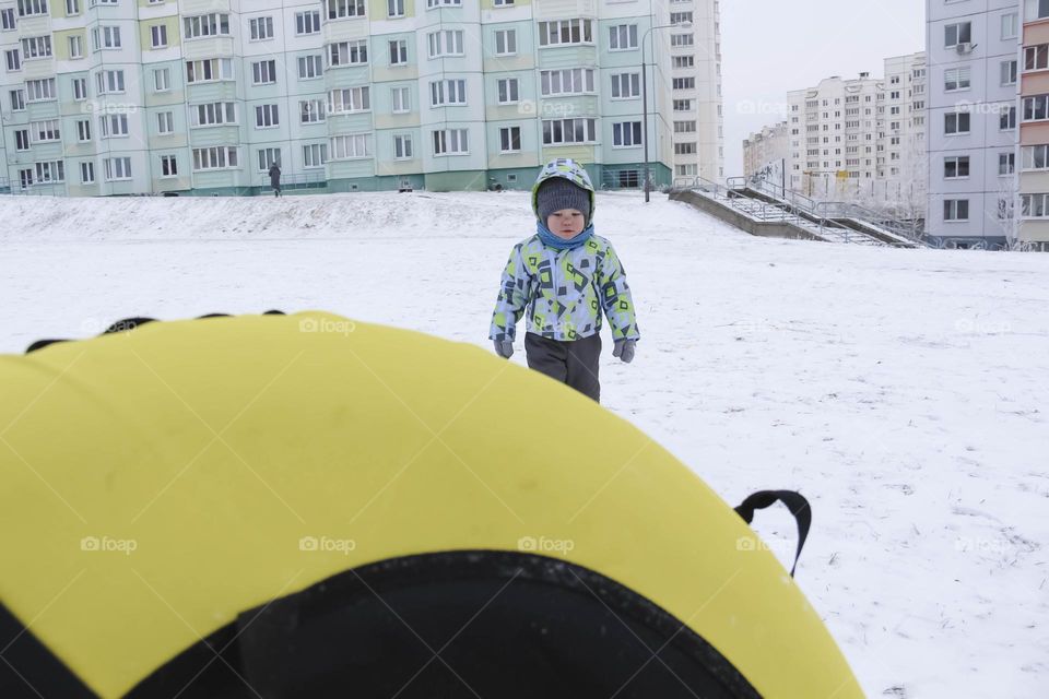 A small, carefree boy walks in the white snow in winter and rides a tubing in the park, near trees in the snow.