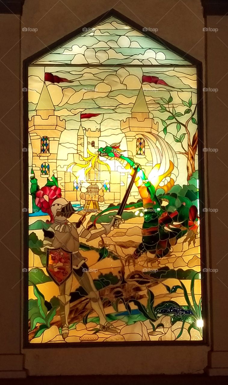 Knight and Dragon scene from a Castle Hotel in rain glass with many golden bright colors shining through this stain glass artwork.