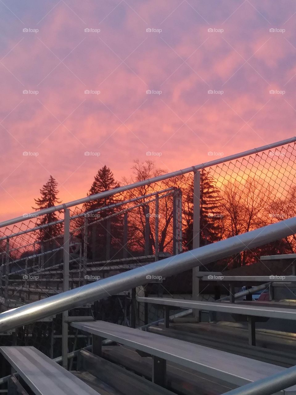 Bench at Brookside west park in Ohio, beautiful sunset sky with rolling clouds and pops of orange yellow pink and purple against the trees silhouette and grey bars of the bleachers 