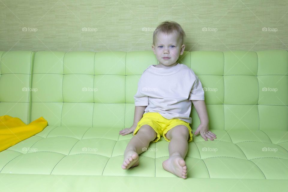 A portrait of a child sitting at home on a green sofa depicts different emotions in yellow shorts.