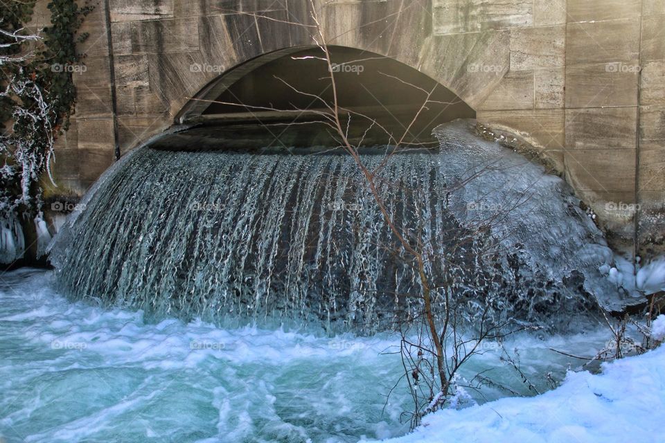 A small waterfall, frozen at the edge, falls under a sandbrown stone bridge into a green river 