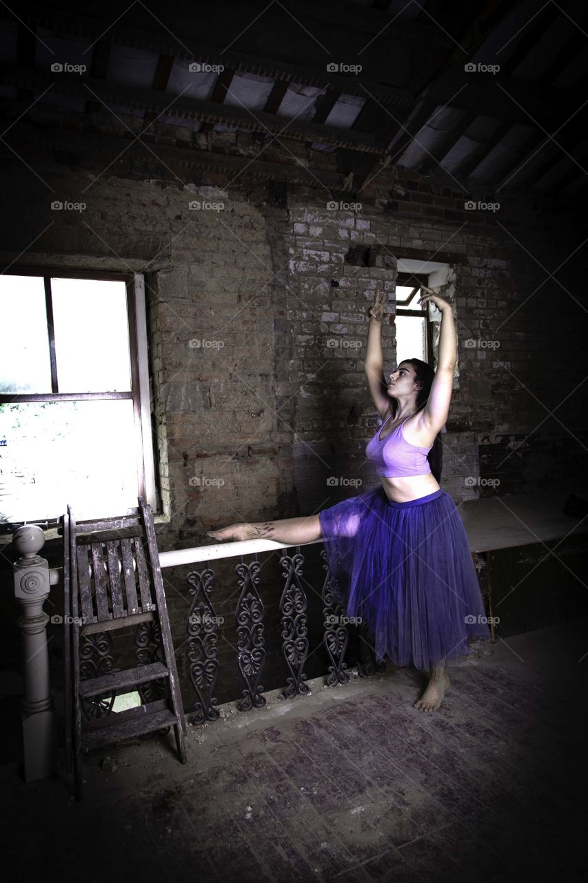 Girl dancing in a derelict building. Leg stretched on top of stair rail at the top of the stairs. Holding a dance pose. Step ladders leaning against hand rail.