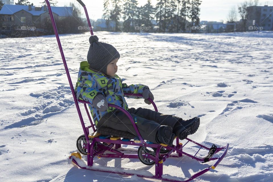 A child in winter in winter jackets, pants, a hat and boots on white snow on the street and in the park in nature plays winter fun and sleds.