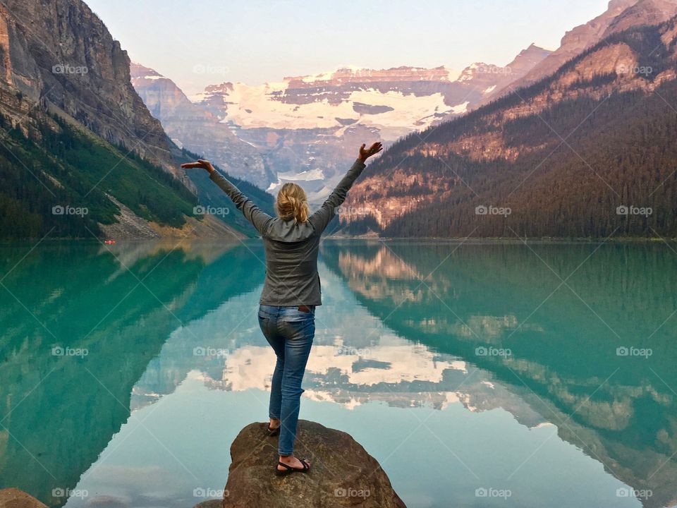 Woman standing on large rock in front of beautiful Lake Louise in Canada's Rocky Mountains with arms outstretched in praise and wonder 