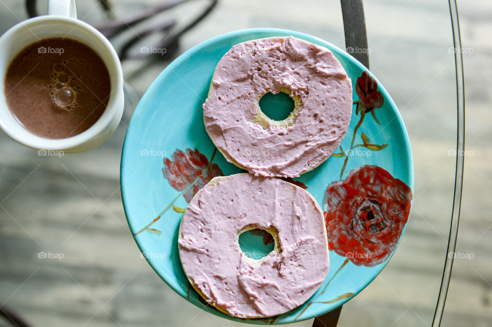 Colorful plate of purple berry cream cheese on an open bagel with a mug of chocolate milk on a glass table