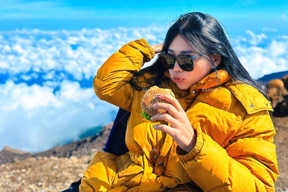 Portrait of young woman enjoying burger on mountain top above beautiful clouds