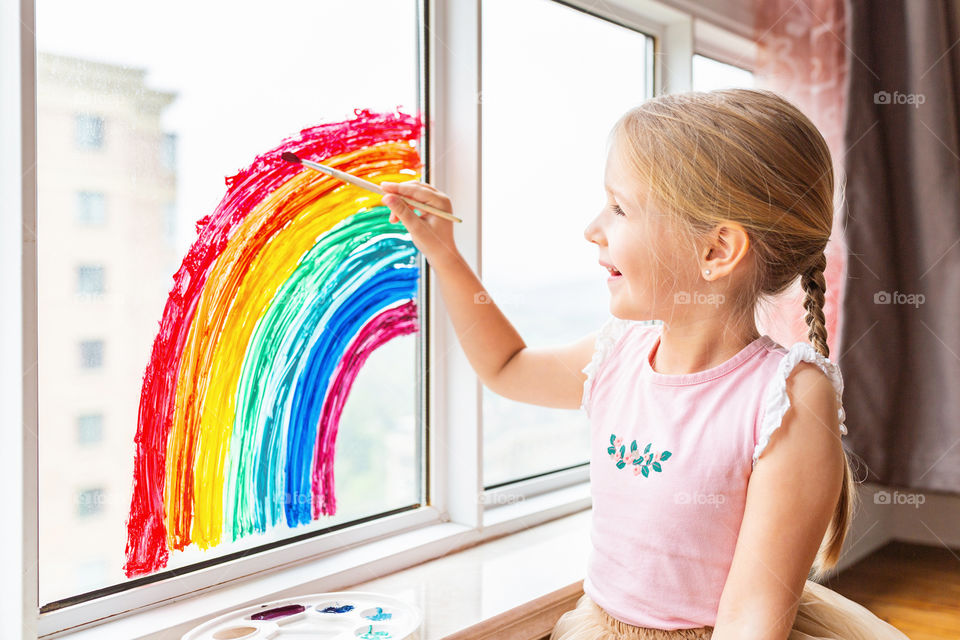 Happy little Caucasian girl with blonde hair painting rainbow during COVID-19 coronavirus quarantine and self isolation 