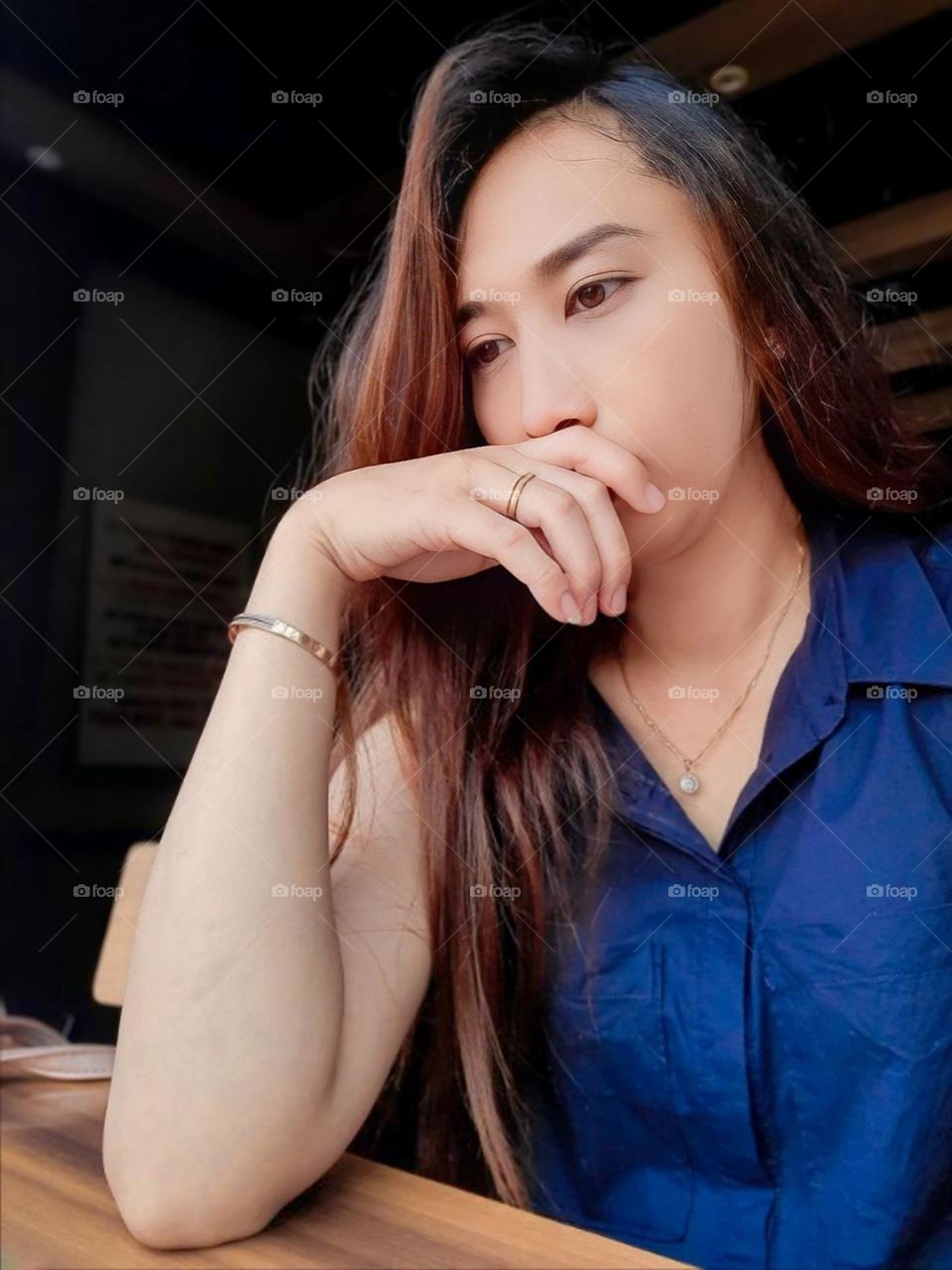 Close-up of a young woman contemplating, wearing a blue shirt and sitting in front of a wooden table. The person looks relaxed and stylish with subtle jewellery