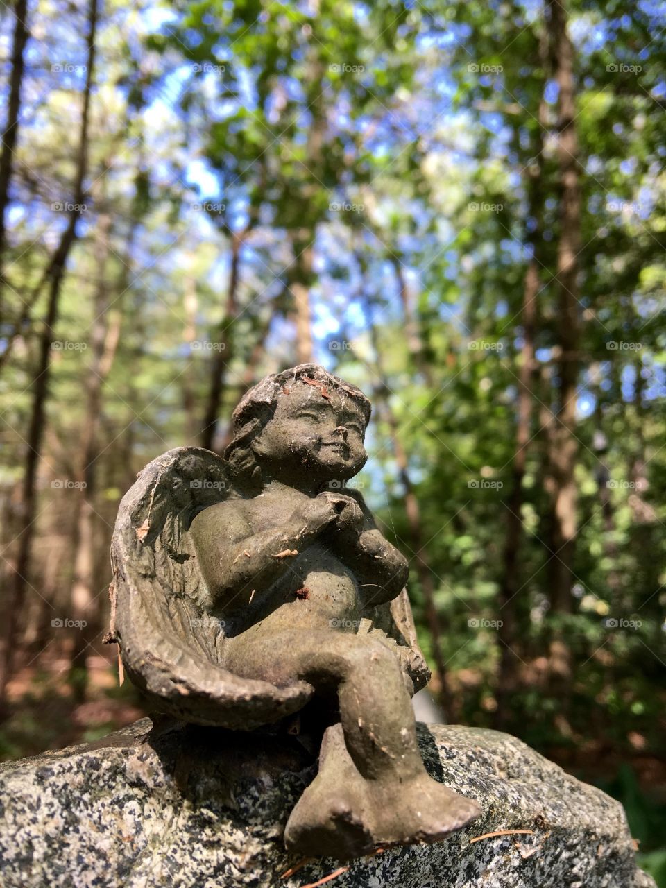 Small Angel sitting on tall rock in the woods, blue sky seen through the trees.