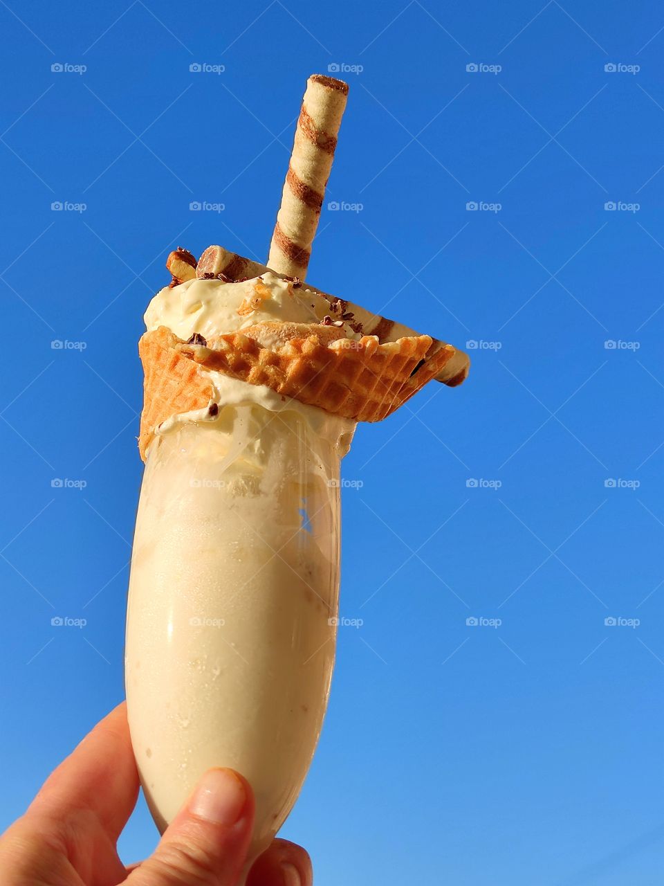 A woman's hand holds a glass of ice cream, the edge of which is wrapped with a waffle ribbon. Ice cream sprinkled with chocolate chips and wafer rolls. Sunny day. Blue sky