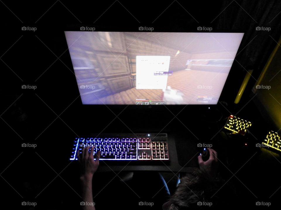 Teenager boy playing Minecraft on the gaming computer seen from a high angle and seeing his hand playing using the keyboard and the mouse accessories in a dark room.