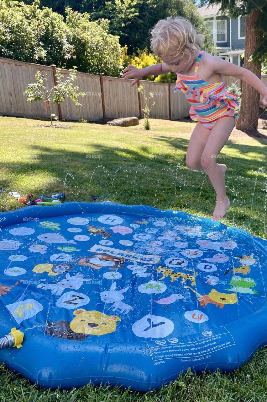 A young child jumps onto a splash pad in the backyard to cool off on a hot summer day