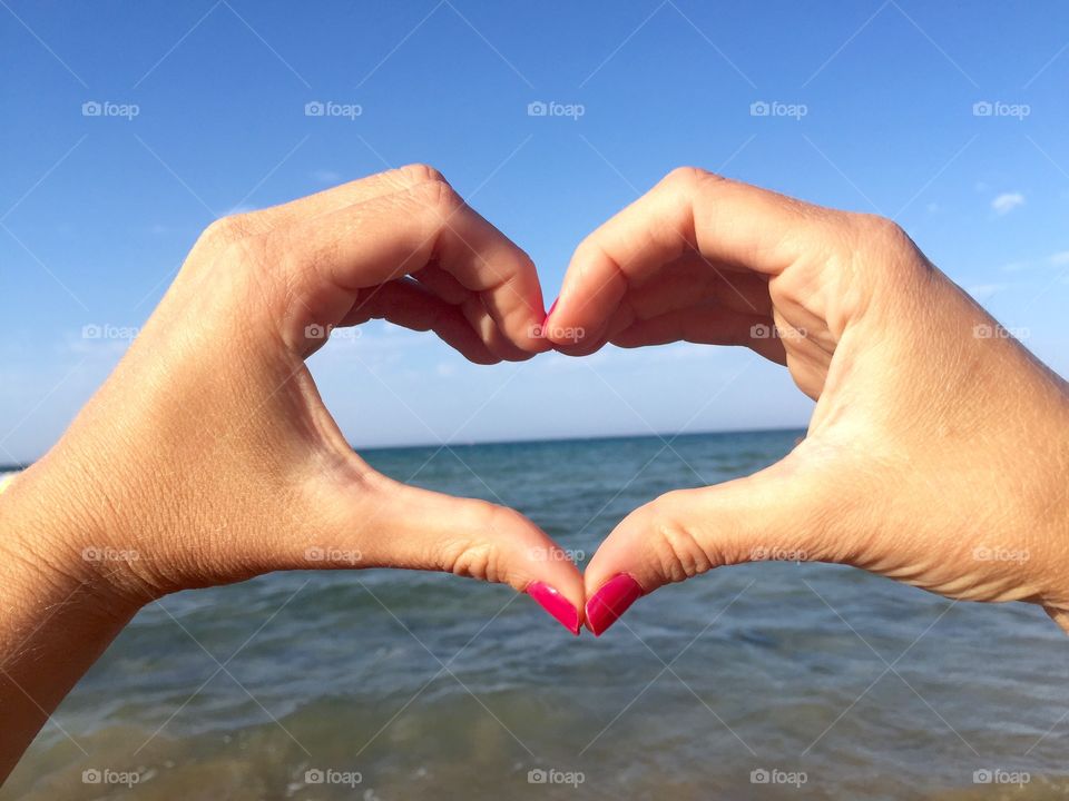Heart made of woman's hands wearing pink nail polish over the sea