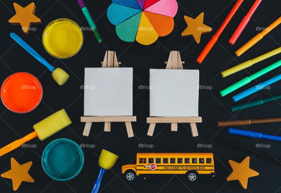 Two small white easels with colorful pencils, felt pens, brushes and paint inks with watercolors and an empty notepad lie on a black background, flat lay close-up.