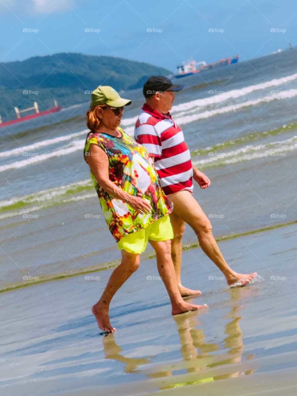 Shot of a Couple hiking in the beach,at Santos, Brasil. Beautiful Reflections in the sand, man is using a striped shirt, both using hats, basebay caps.