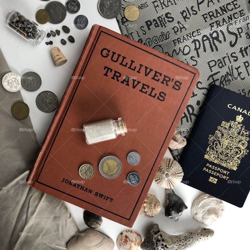 A vintage book of Gullivers travels and surrounded by articles of vacation and travel such as money, shells, a passport, and sand