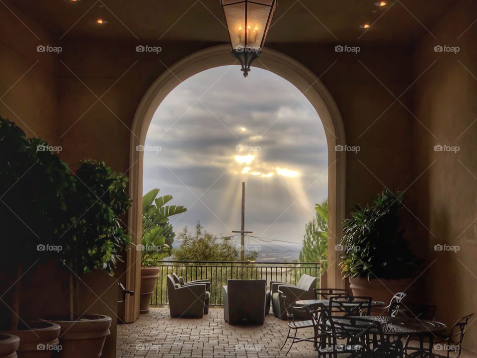Peering out from a balcony with the sun shining through the clouds with chairs placed in view and trees in the outer frame. A cross in center frame.