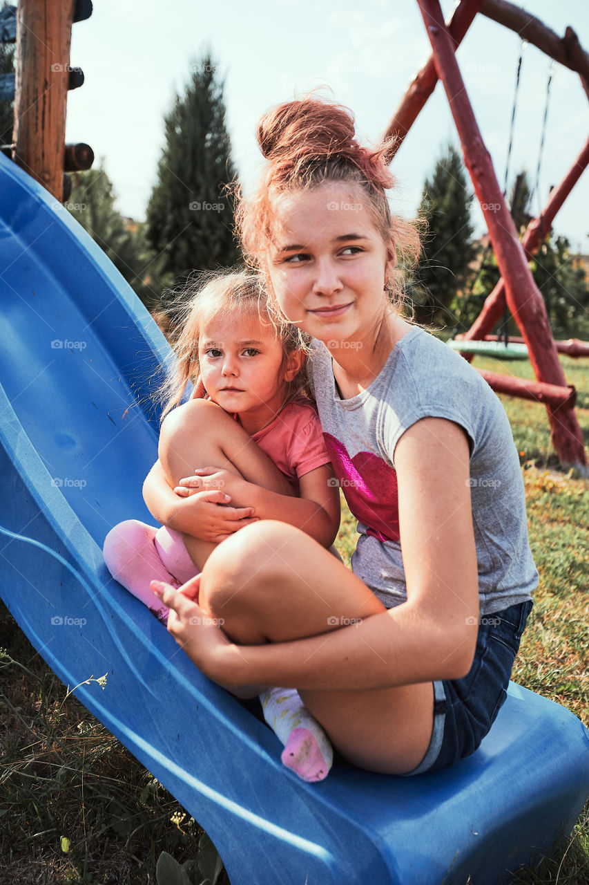 Teenage girl playing with her younger sister in a home playground in a backyard. Happy smiling sisters having fun on a slide together on summer day. Real people, authentic situations