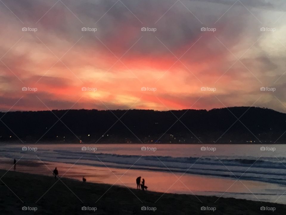 A beautiful Monterey sunset allures people to walk along the beach to enjoy her splendor. A couple walks their dog while other people stand looking out at the multicolored sky and ocean, peacefully taking in all the immeasurable beauty. 