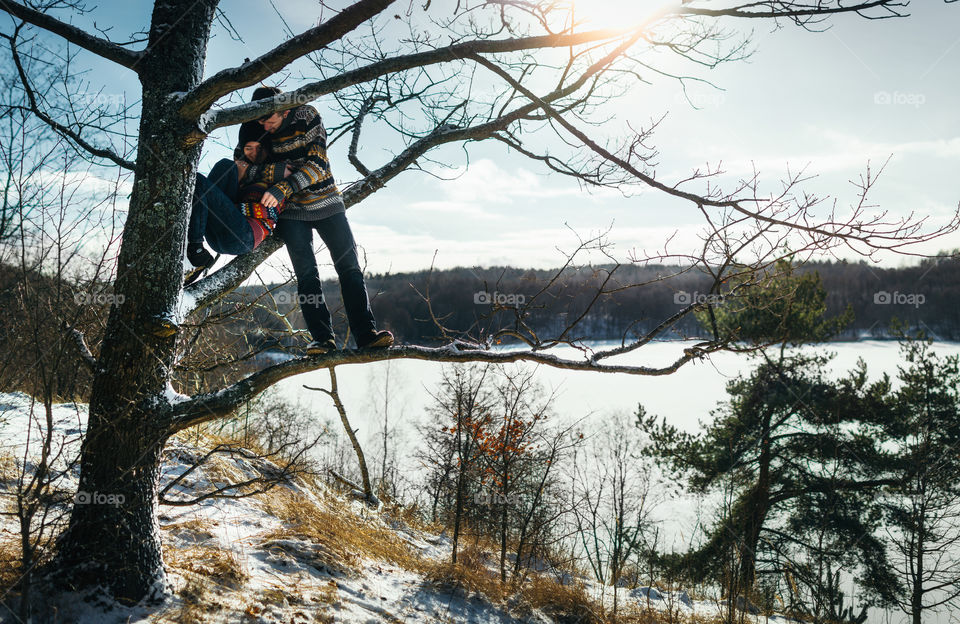 Joyful cute couple embraces. Young man in a sweater hugs a girlfriend sitting on tree branch in winter.