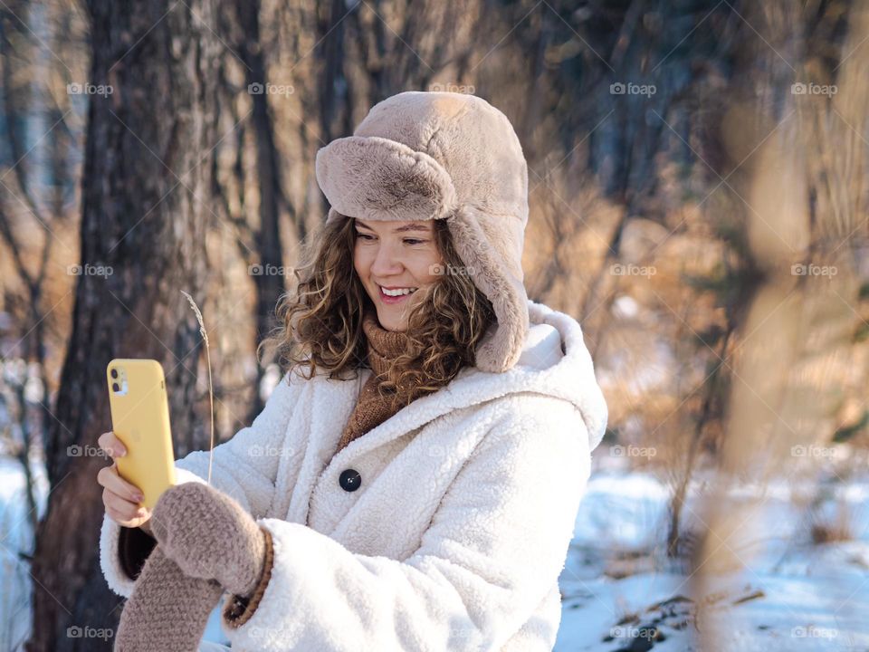 Young beautiful woman with curly hair in fur coat and hat with ear flaps taking selfie in winter forest