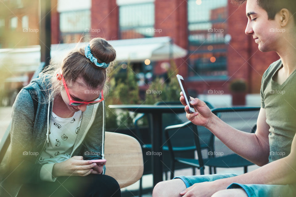 Couple of friends, teenage girl and boy, having fun using smartphones sitting in center of town, spending time together