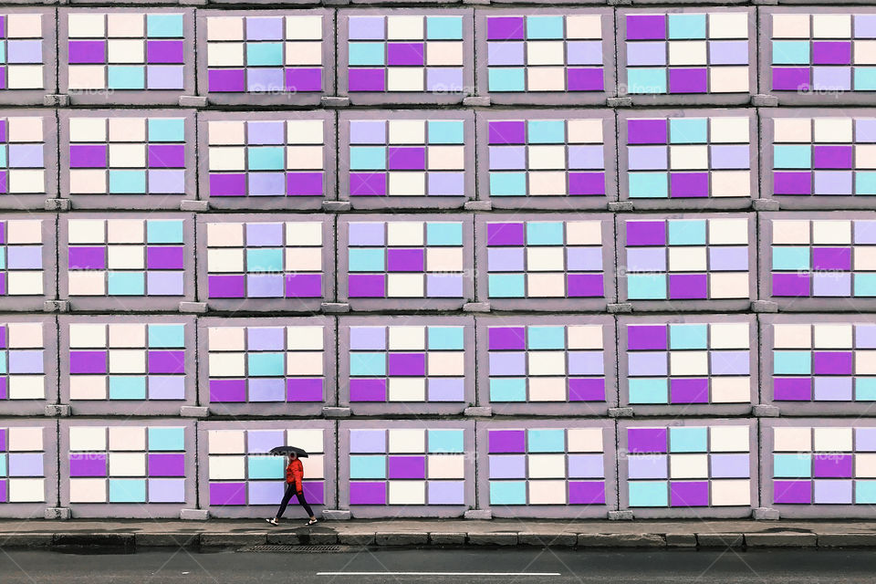 Tiny human with umbrella walking in front of a colorful geometric wall 