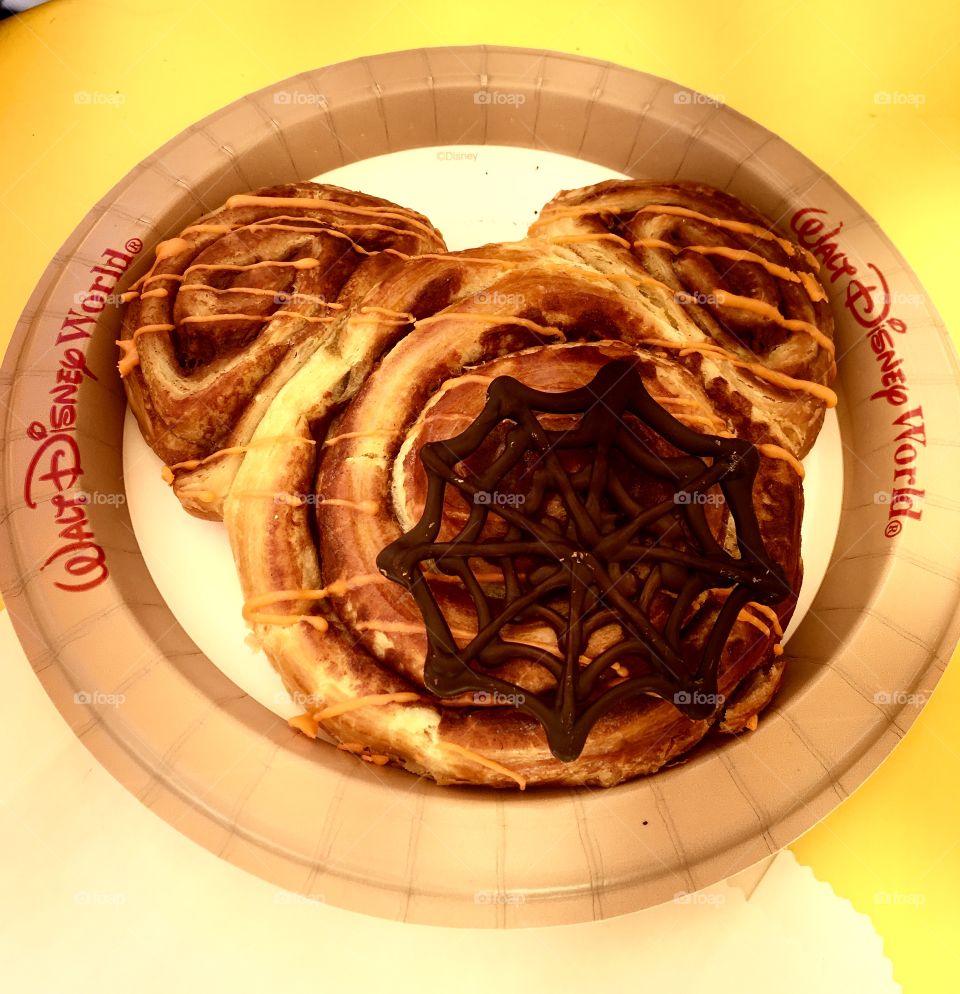 A Mickey Mouse shaped cinnamon bun with a chocolate spider web candy on top sitting on a paper plate on a yellow table. Taken in Disney World during Halloween 