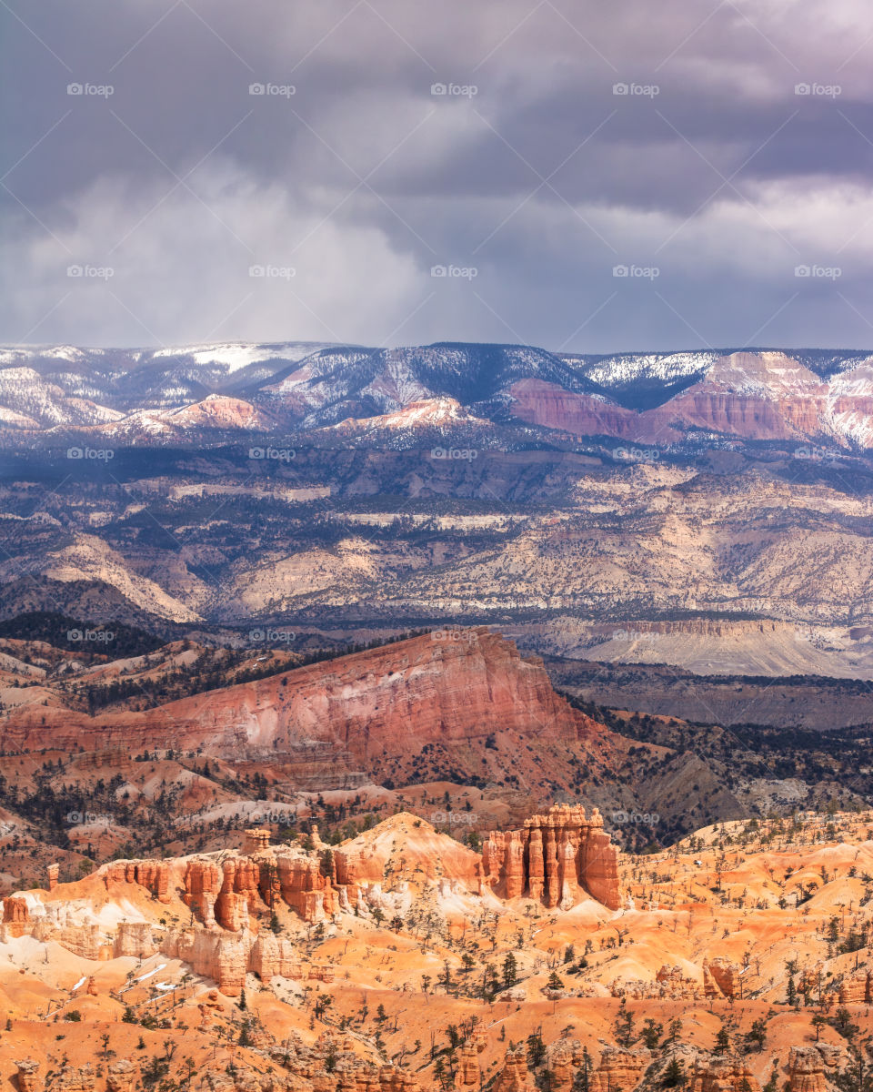 Layers of unique rock formations and mountains with shadows and golden sunlight on them, under a dark moody sky. Bryce Canyon National Park. 